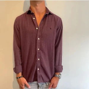 Mörklila skjorta med knappar - Säljer en stilren mörklila skjorta med klassisk krage och vita knappar. Skjortan har lång ärm och är tillverkad i ett mjukt bomullstyg. Perfekt för dig som gillar en enkel men snygg look.