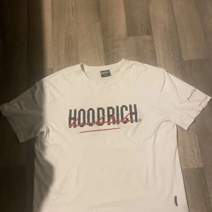 Vit HOODRICH t-shirt med tryck - Vit t-shirt från HOODRICH i storlek Large. Snyggt tryck med HOODRICH-logga i svart och rött både fram och bak. Rund halsringning och korta ärmar. Skön och klassisk passform i mjuk bomull. Extra detaljer med text på ena ärmen.Storlek: L 	•	Färg: Vit med svart och röd text/design 	•	Skick: Ny och fin 	•	Beskrivning: Snygg original Hoodrich T-shirt med tryck både fram och bak. Framtryck med tydlig “HOODRICH”-logga och röd handskriven effekt. Litet patch-märke nedtill. Perfekt passform för en avslap