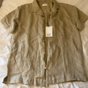 Beige linneskjorta These Glory Days - Snygg beige skjorta från These Glory Days i storlek S. Skjortan är tillverkad i linne, har korta ärmar, klassisk krage, bröstficka och knäppning framtill. Perfekt för en chill och stilren look under varmare dagar.