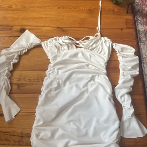 Vit kort satinklänning med puffärmar - Supersnygg vit kort klänning i satin med avtagbara puffärmar och knytning vid halsen. Klänningen har rynkade detaljer längs sidorna och en lätt v-ringad urringning. Perfekt för dig som vill sticka ut med en trendig look.    Priset+frakt