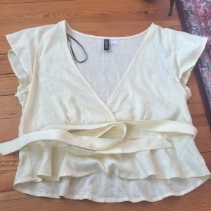 Ljusgul omlottopp från Gina Tricot - Säljer en ljusgul omlottopp från Gina Tricot med korta ärmar och knytband i midjan. Toppen har v-ringning och volangdetalj nedtill, perfekt för en fräsch och somrig look. Priset + frakt