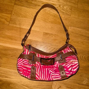 Vintage väska  - Cool axelväska i röd- och vit zebramönstrad textil med bruna skinnliknande detaljer och bred axelrem. Väskan har snygga spännen, dragkedja upptill och en praktisk ficka med dragkedja på baksidan. Perfekt för dig som vill sticka ut med din stil.