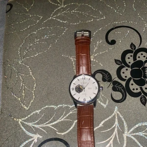 Paterson automatisk klocka brun läder - Snygg automatisk klocka från Paterson med vit urtavla och öppet hjärta. Klockan har ett klassiskt runt silverfärgat boett och ett brunt läderarmband med krokodilpräglat mönster. Kommer i original ask med guldtext. Perfekt för dig som gillar stilrena accessoarer.