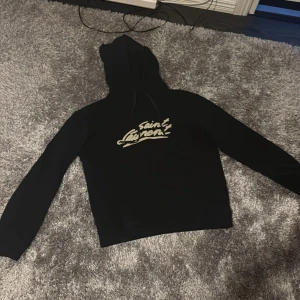 Saint Laurent hoodie - Svart hoodie från Saint Laurent med stort beige logotyptryck på bröstet. Klassisk modell med huva och snörning, samt långa ärmar. Perfekt för dig som gillar streetstyle och vill ha en snygg statementtröja.