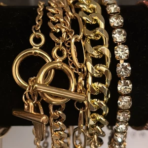 Guldiga armband med kedjor och stenar - Flera snygga armband i guldton, vissa med chunky kedjor och andra med glittrande stenar. Perfekt för att mixa och matcha för en cool och trendig look. Armbanden har olika länkar och detaljer som ger en lyxig känsla. 5st