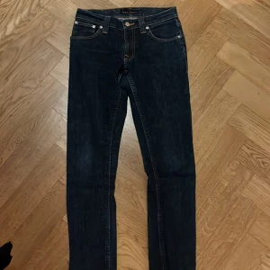 Mörkblå jeans från Nudie Jeans - Snygga mörkblå jeans från Nudie Jeans med klassisk femficksdesign och kontrastsömmar. Byxorna har raka ben och ikonisk Nudie-logga på bakfickorna samt läderpatch i midjan. Tillverkade i slitstark bomullsdenim för en tidlös look.