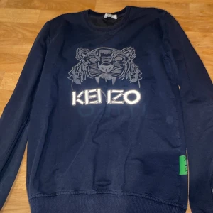 Mörkblå Kenzo sweatshirt med tiger - Säljer en mörkblå sweatshirt från Kenzo med det ikoniska tigermotivet och broderad logga på bröstet. Tröjan har ribbade muddar, rund hals och en grön Kenzo-detalj i sidan. Perfekt för dig som gillar streetwear och vill sticka ut.