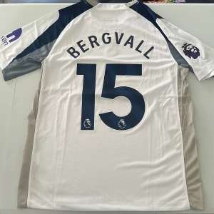 Tottenham Bergvall 15 matchtröja Nike - Säljer en Tottenham Hotspur fotbollströja med Bergvall 15 på ryggen. Tröjan är vit med marinblå och grå detaljer, röd AIA-logga på bröstet och Premier League-märke på ärmen. Tillverkad i lätt och ventilerande material, perfekt för match eller träning. Replika, endast handtvätt gäller!