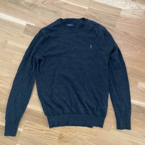Mörkgrå merinoullströja från Polo Ralph Lauren - Snygg mörkgrå tröja från Polo Ralph Lauren i tvättbar merinoull. Slim fit-modell med rund halsringning och broderad logga på bröstet. Långärmad och perfekt för lager-på-lager-stil under höst och vinter.