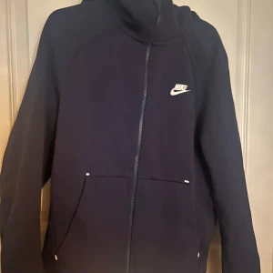 blå Nike hoodie med dragkedja - Säljer en blå hoodie från Nike med vit logga på bröstet. Tröjan har hel dragkedja, huva och två fickor framtill. Perfekt för chill dagar eller när du vill ha en sportig look. Materialet är mjukt och skönt, troligtvis bomullsmix.