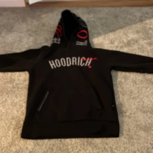 Svart HOODRICH hoodie med tryck - Svart hoodie från Hoodrich med stor vit logga på bröstet och röda detaljer,fickor med dragkedjor, tryck på luvan och på ryggen. Knappt andvänd och inga synliga tecken andvändning och nypris är 899kr.