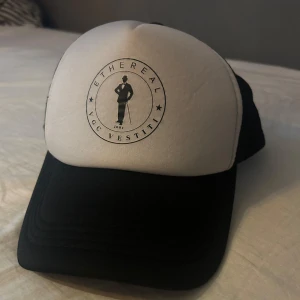 Svartvit truckerkeps Ethereal VGC Vestiti - Snygg truckerkeps i svart och vitt från Ethereal VGC Vestiti. Framsidan är vit med svart tryck och logga, medan baksidan är i svart mesh med justerbar knäppning. Perfekt accessoar för en cool och avslappnad stil.