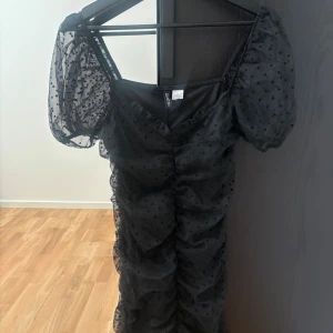 Svart prickig meshklänning med puffärm - Supersöt svart kortklänning från Boohoo i mesh med prickigt mönster. Klänningen har puffiga korta spets ärmar, rynkade detaljer längs hela kroppen och v-ringning. Perfekt för dig som gillar trendiga och lekfulla plagg!