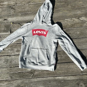 Grå hoodie från Levi's med logga - Säljer en grå hoodie från Levi's med stor röd logga på bröstet. Tröjan har huva, magficka och långa ärmar. Perfekt för dig som gillar en avslappnad och sportig stil.