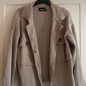 Beige stickad kofta från Pieces - Säljer en beige stickad kofta från Pieces i storlek M. Koftan har ribbad struktur, stora fickor framtill med knappar och en avslappnad passform, storlek M men jag tycker att den är större än så