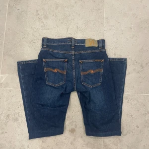 Mörkblå jeans från Nudie Jeans - Nya! Snygga mörkblå jeans från Nudie Jeans med klassisk femficksdesign och orange kontrastsömmar. Byxorna har raka ben och normal passform, tillverkade i slitstarkt denim med Nudie-logga på bakfickan. Perfekta för dig som gillar stilrena och tidlösa jeans.