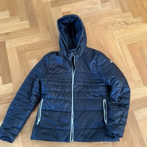 Svart pufferjacka från Adidas L - Svart täckjacka från Adidas med huva och quiltade detaljer. Jackan har dragkedja framtill och två fickor med dragkedja. Adidas-logga på ärmen. Perfekt för kyliga dagar och har en sportig vibe.