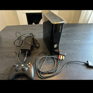 Xbox 360 Slim - Xbox 360 Slim i svart med original strömadapter, AV-kabel och en trådbunden handkontroll. Konsolen och tillbehören är i gott begagnat skick med mindre ytliga repor. Perfekt för dig som vill spela klassiska Xbox 360-spel direkt!