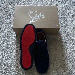 Christian Louboutin  - Exklusiva marinblå sneakers från Christian Louboutin med klassisk röd sula och snygg snörning. Skorna har en stilren design i mocka och kommer med originalkartong, dustbag och äkthetsbevis. Perfekta för dig som vill sticka ut med lyxiga detaljer.