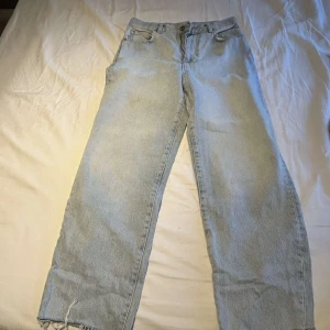 Ljusblå raka jeans med rå kant - Säljer ett par ljusblå jeans med rak passform och rå, fransig kant nertill. Klassisk femficksmodell med knapp och dragkedja framtill. Perfekta för en avslappnad och trendig look.