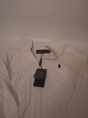 Vit stickad half zip tröja Polo Ralph Lauren - Säljer en vit stickad tröja från Polo Ralph Lauren med half zip och kabelstickat mönster. Tröjan har en ribbad krage och det klassiska broderade logot på bröstet. Perfekt för dig som gillar stilrena och tidlösa plagg.