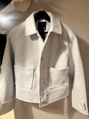 Ljusgrå overshirt från 157, S - Snygg ljusgrå overshirt från 157 i storlek S. Jackan har klassisk krage, stora fickor framtill och knappar längs hela framsidan. Materialet är mjukt och känns som ullmix, perfekt för lager-på-lager. Enkel och clean design som är lätt att matcha. Aldrig använd, inköpt för 400kr