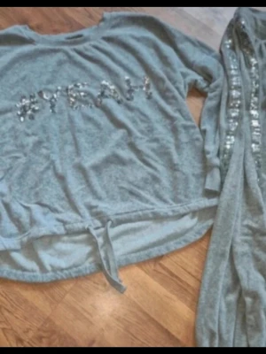 Grå mjukisbyxor och tröja med paljettdetalj Esmara storlek L/XL - Grå mjukisbyxor från Esmara med bred mudd i benslut och en snygg rad med silverfärgade paljetter längs sidorna. Byxorna är i mjukt material och har en relaxed passform, perfekta för chill dagar hemma. Snygg matchande tröja till paljettdetaljer. Velourmaterial. 