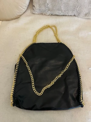 Stor svart Stella mccartney liknade väska - Snygg svart lite större handväska med kraftiga guldfärgade kedjor som axelrem och dekor längs kanterna. Väskan har ett stort huvudfack och en innerficka med dragkedja.💖 väskan har aldrig använts, så säljer pågrund av att den inte kommer till användning