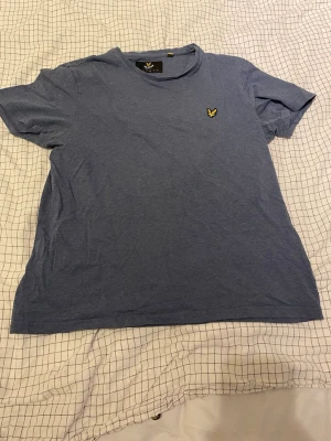 Blå t-shirt från Lyle & Scott - Säljer en blå t-shirt från Lyle & Scott med klassisk rund hals och korta ärmar. T-shirten har en gul broderad logga på bröstet och är gjord i mjuk bomull. Perfekt för dig som gillar enkel och stilren design.