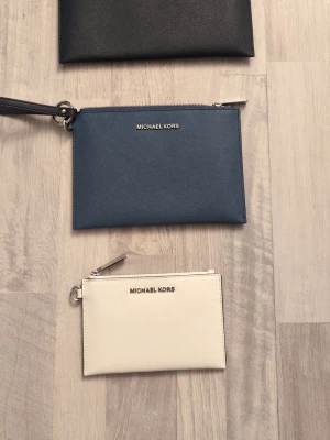 Michael Kors kuvertväskor i skinn - Tre stilrena kuvertväskor från Michael Kors i skinn. En är svart, en marinblå med handledsrem och en är vit. Alla har silverfärgad dragkedja och logga framtill. Perfekt storlek för det viktigaste och snygg att bära i handen. Samtliga 3st för 590 alternativt så är styckpris för den stora mörkblåa 280, mellan den blåa 240 kr den lilla vita 140kr