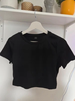 Svart croppad topp från SHEIN - Säljer en svart croppad topp från SHEIN i storlek M. Toppen har rund halsringning och korta ärmar, tillverkad i mjukt syntetmaterial. Enkel och stilren design som är lätt att matcha med olika outfits.