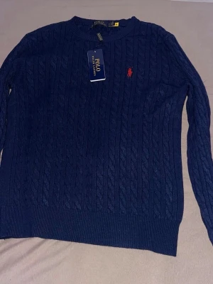 Blå stickad tröja från Polo Ralph Lauren - Säljer en marinblå stickad tröja från Polo Ralph Lauren med klassiskt kabelmönster och rund hals. Tröjan har lång ärm och en röd broderad logga på bröstet. Perfekt för dig som gillar stilrena och tidlösa plagg.