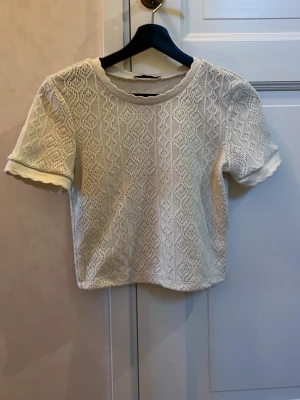 Vit spetsig topp från Zara - Säljer en söt vit topp från Zara i storlek S. Toppen har ett spetsigt stickat mönster och korta ärmar med vågiga kanter. Rund halsringning och croppad passform ger en fräsch och ungdomlig vibe. Perfekt för vår och sommar!