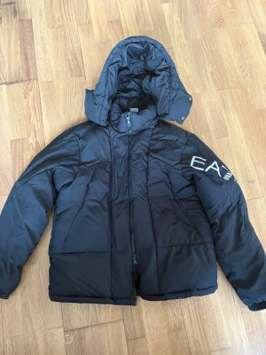 Svart pufferjacka från Emporio Armani - Säljer en svart pufferjacka från Emporio Armani med stor logga på ärmen. Jackan har huva, dragkedja framtill och en riktigt skön, vadderad känsla. Perfekt för kalla dagar och ger en clean streetwear-look.