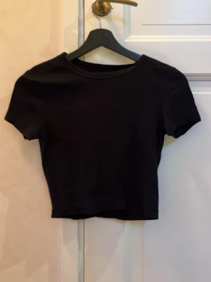 Svart ribbad croppad topp Even&Odd - Säljer en svart croppad topp från Even&Odd. Toppen har korta ärmar, rund halsringning och är ribbad i materialet vilket ger en snygg struktur. Perfekt till jeans eller kjol för en enkel men stilren look.