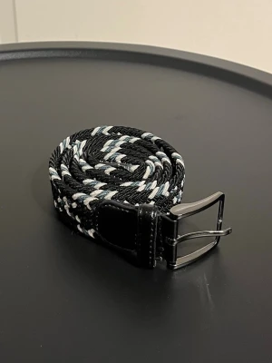 Flätad bälte 105 cm - Snyggt flätat bälte i svart, vitt och lite ljusblått. 105 cm långt, men går att stretcha ut. Bältet har ett rutigt mönster och detaljer i svart skinn vid spännet och änden. Metallspänne i silverfärg ger en clean look. Perfekt accessoar för att lyfta din outfit. Använt några gånger, men med omsorg. 