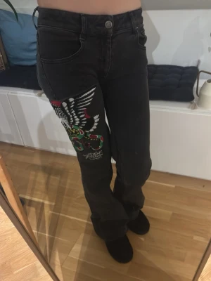 Svarta Ed Hardy jeans med broderi - Svarta jeans från Ed Hardy med stort broderat motiv av en örn och en färgglad orm på ena benet. Snyggt Ed Hardy-tryck på bakfickan. Modellen är bootcut och normal passform. om sydliggrenen för med låg midjad.