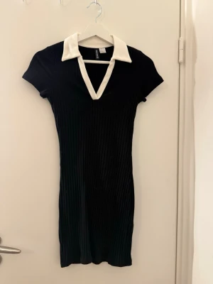 Svart ribbad kortklänning från H&M - Säljer en svart ribbad kortklänning från H&M Divided i storlek XS. Klänningen har vit krage och v-ringning, samt korta ärmar. Materialet är mjukt och stretchigt, perfekt för en cool och trendig look. Passformen är slim och klänningen har en klassisk, stilren design.