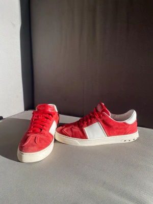 Valentino flycrews röda sneakers - Säljer ett par Valentino flycrews sneakers i rött mocka och vitt skinn. Skorna har röda snören, vit sula med coola detaljer och en kontrasterande vit panel på sidan. Perfekta för dig som vill sticka ut med en lyxig och sportig vibe.