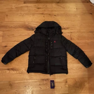 Svart pufferjacka från Polo Ralph Lauren - Svart pufferjacka från Polo Ralph Lauren med huva och två dragkedjeförsedda fickor. Jackan har det klassiska röda Polo-logot broderat på bröstet och quiltad design.