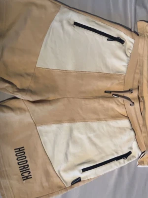 Beige och vita shorts från Hoodrich - Säljer ett par snygga Hoodrich shorts i beige och vitt med svarta dragkedjor på fickorna och snörning i midjan. De har en cool paneldesign och Hoodrich-logga på benet. Perfekta för chill och streetwear. Materialet är mjukt och känns som bomull.