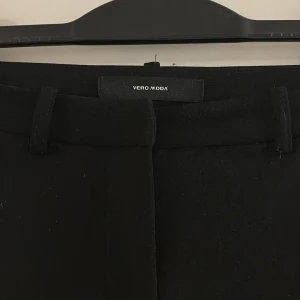 Svarta kostymbyxor från Vero Moda - Snygga svarta kostymbyxor från Vero Moda med raka ben och klassiska bälteshällor. Byxorna har en stilren design och är tillverkade i ett mjukt material som ger en skön känsla. Perfekta för dig som gillar en clean och enkel look.