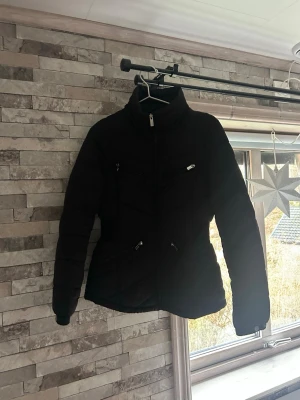 Svart pufferjacka från Livelo - Svart pufferjacka från Livelo med hög krage och fyra snygga dragkedjor framtill. Jackan har quiltad design och långärmade ärmar. Perfekt för dig som vill ha en stilren och varm jacka med coola detaljer.