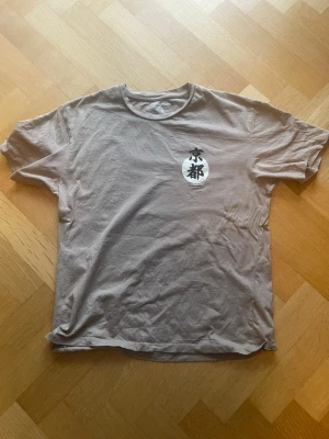 Beige Kyoto t-shirt från Jack & Jones - Säljer en beige t-shirt från Jack & Jones med coolt Kyoto-tryck fram och stort motiv med koi-fiskar och japanska tecken på ryggen. Rund hals och korta ärmar, tillverkad i mjuk bomull. Perfekt för dig som gillar streetstyle och asiatiska influenser.