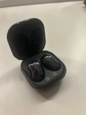 Samsung Galaxy Buds Live - Samsung Galaxy Buds Live trådlösa hörlurar i svart med laddningsetui. Unik bönformad design för bekväm passform och bra ljud. Etuiet har synliga repor men hörlurarna ser ut att vara i gott skick. Perfekt för musik, samtal och träning.