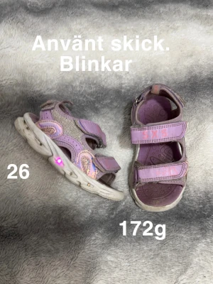 Lila blinkande sandaler med kardborre - Säljer ett par lila sandaler med blinkande sula och dubbla kardborreband. Skorna har meshdetaljer på sidorna och är lätta att ta på. Perfekta för sommarens äventyr 🌸 Sandaler som blinkar. Ej tvättade. Smuts på kardborrebandet och sandalen. Storlek 26