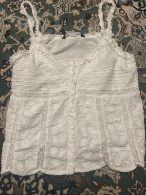 Vit spetsdetaljerad linnetopp från Zara - Supersöt vit linnetopp från Zara med broderade blommor och spetsdetaljer. Toppen har smala axelband, v-ringad hals och dekorativa volanger längs kanterna. Materialet är lätt och luftigt, perfekt för varma dagar. Säljer då jag rensar garderoben!