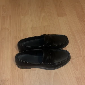 Svarta loafers från Zara i skinnimitation - Säljer ett par klassiska svarta loafers från Zara i skinnimitation. De har en stilren design med rund tå och platt sula, perfekta till både jeans och kostymbyxor. Kommer i originalkartong.
