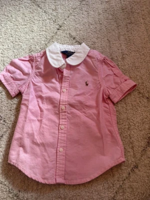 Rosa skjorta från Polo Ralph Lauren - Söt rosa skjorta från Polo Ralph Lauren med vit krage och korta ärmar. Skjortan har knappar framtill och en liten broderad logga på bröstet. Storlek 3 yrs. Helt nytt skick.