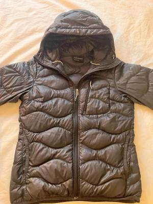 Peak Performance svart dunjacka - Svart dunjacka från Peak Performance i storlek S. Jackan har vågformat quiltat mönster, huva med snörning och dragkedja framtill samt på bröstfickan. Perfekt för kalla dagar och riktigt snygg passform.💓